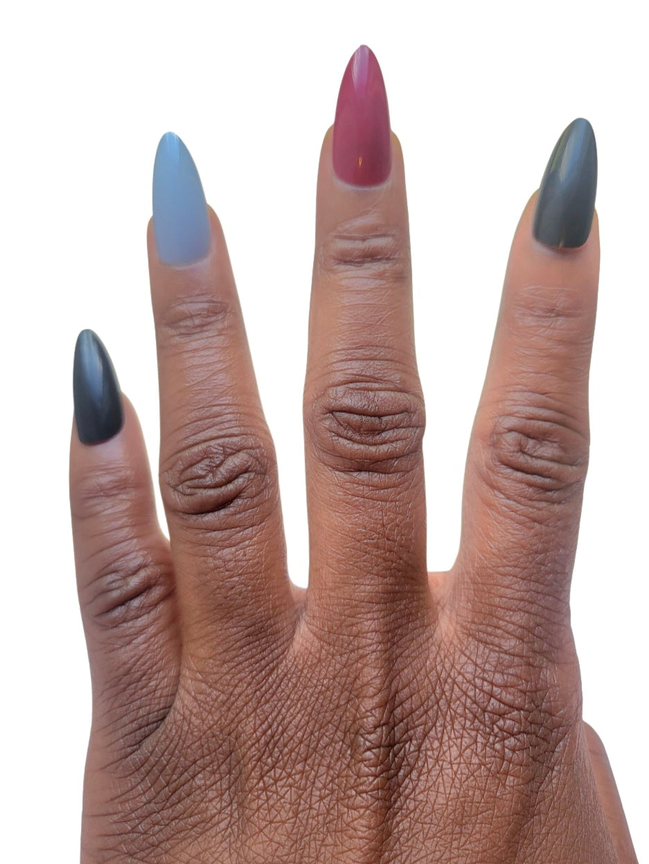 A hand model showcasing the Essentials Press-On Kit by Bloom Gloss Co, featuring matte almond-shaped fake nails in a rich palette of midnight black, muted slate gray, deep wine berry red, and dark olive green.