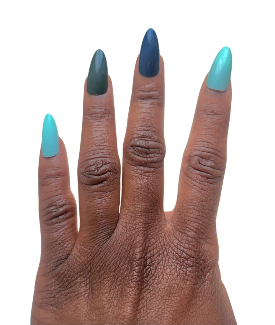 A hand model showcasing the Essentials Press-On Kit by Bloom Gloss Co, wearing glossy almond-shaped fake nails in a mix of deep slate, moss green, and mint sky pastel tones.