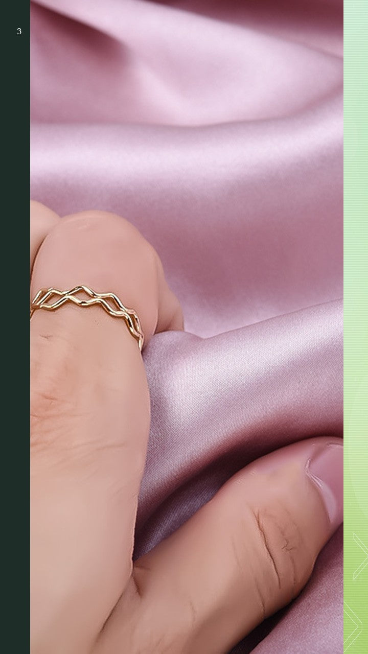 A close-up view of the Ripple Ring by Bloom Gloss Co worn on a hand model, showcasing the 18K gold plated wavy zigzag geometric pattern against a soft pink silk fabric background.